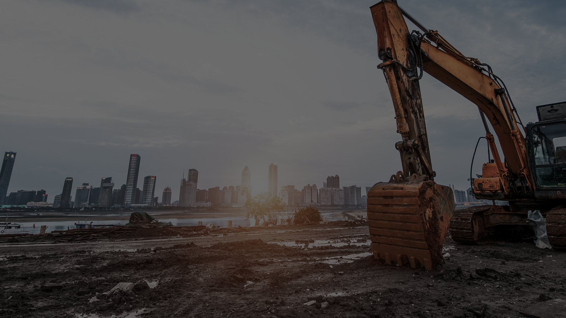 Construction excavator on a muddy jobsite with a city skyline at dawn